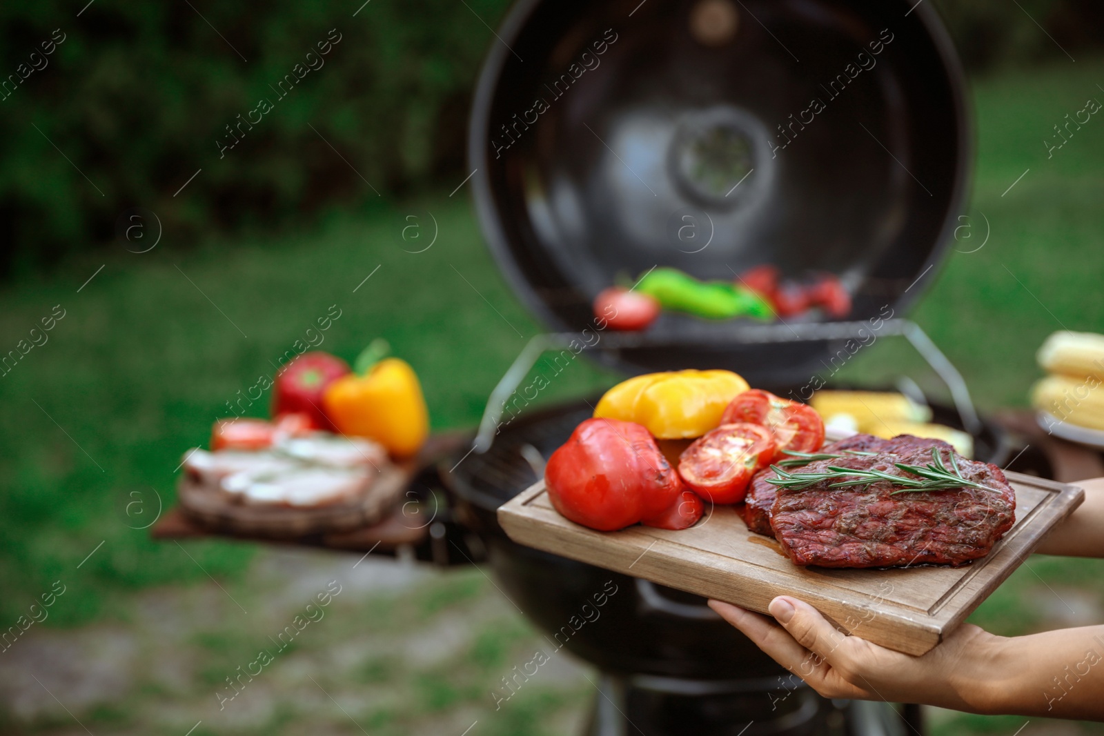 Photo of Woman holding wooden board with grilled vegetables and steak outdoors, closeup. Summer barbecue
