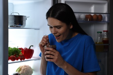 Young woman eating sweet jam near modern refrigerator at night