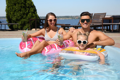 Happy family with inflatable rings and cocktails in outdoor swimming pool on sunny summer day