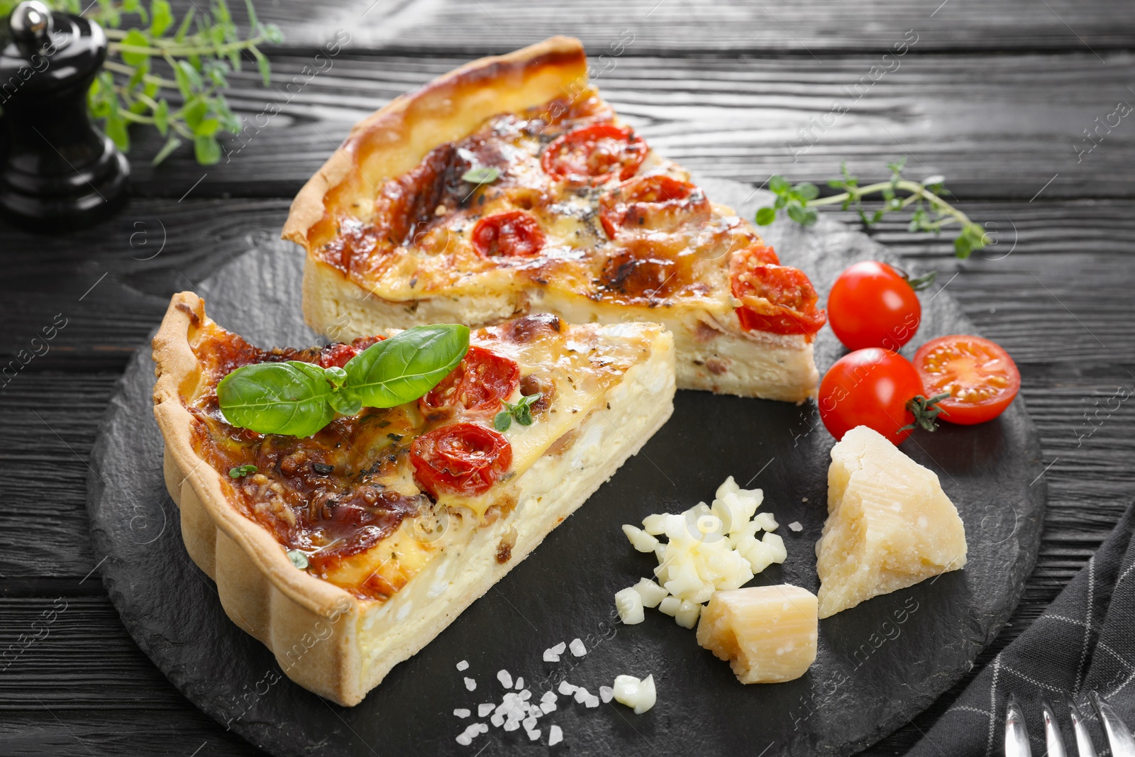 Photo of Pieces of delicious homemade quiche with prosciutto and ingredients on black wooden table, closeup