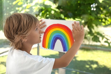 Little boy holding rainbow painting near window. Stay at home concept