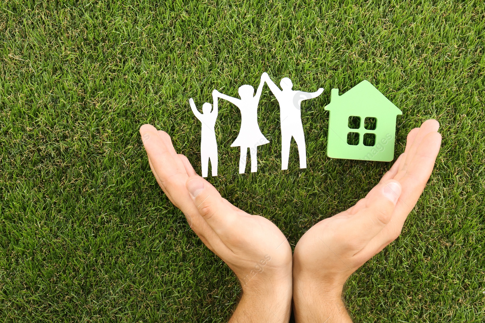 Photo of Man holding hands near figures of house and family on green grass, top view