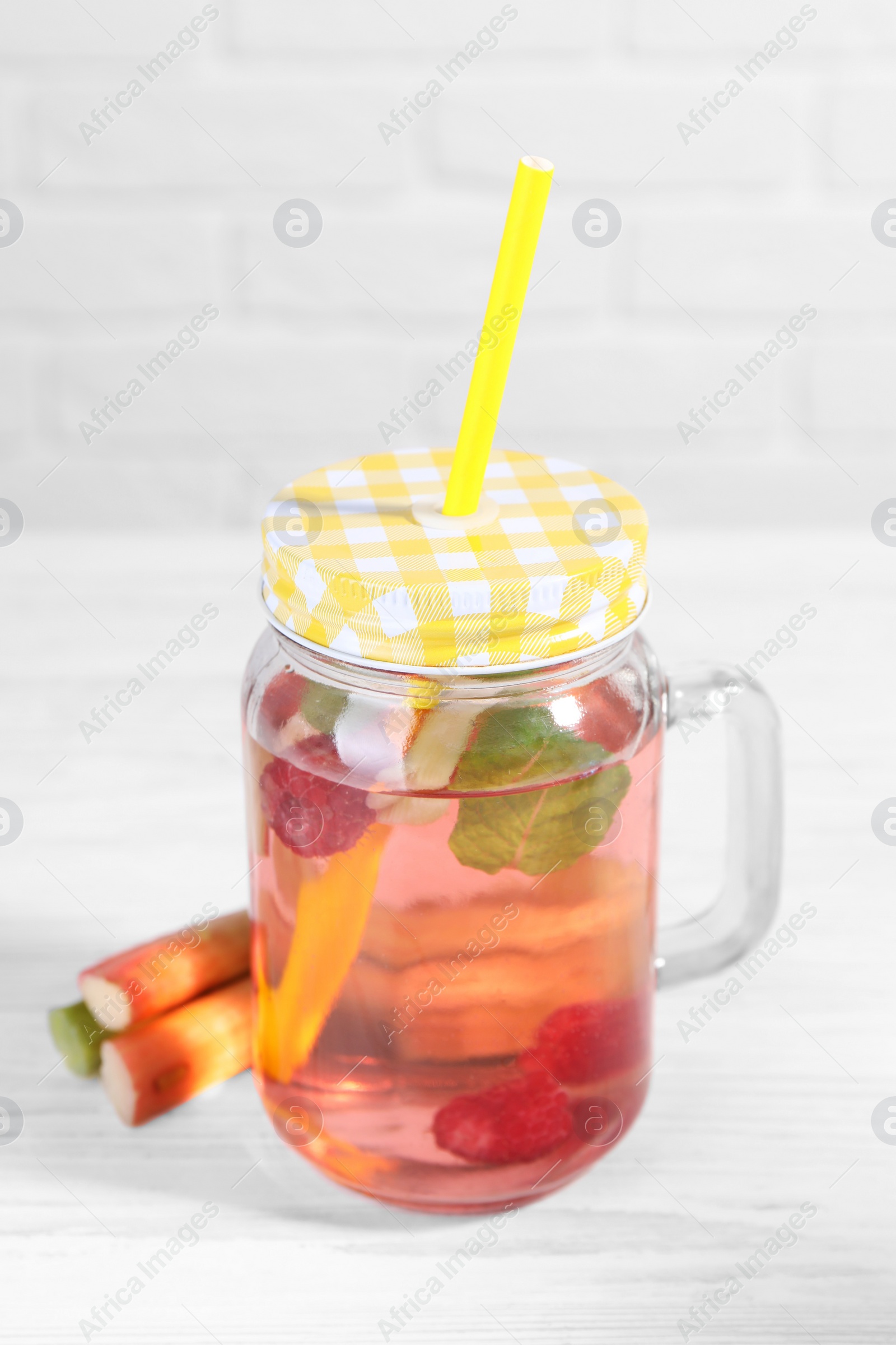 Photo of Mason jar of tasty rhubarb cocktail with raspberry and stalks on white wooden table, closeup