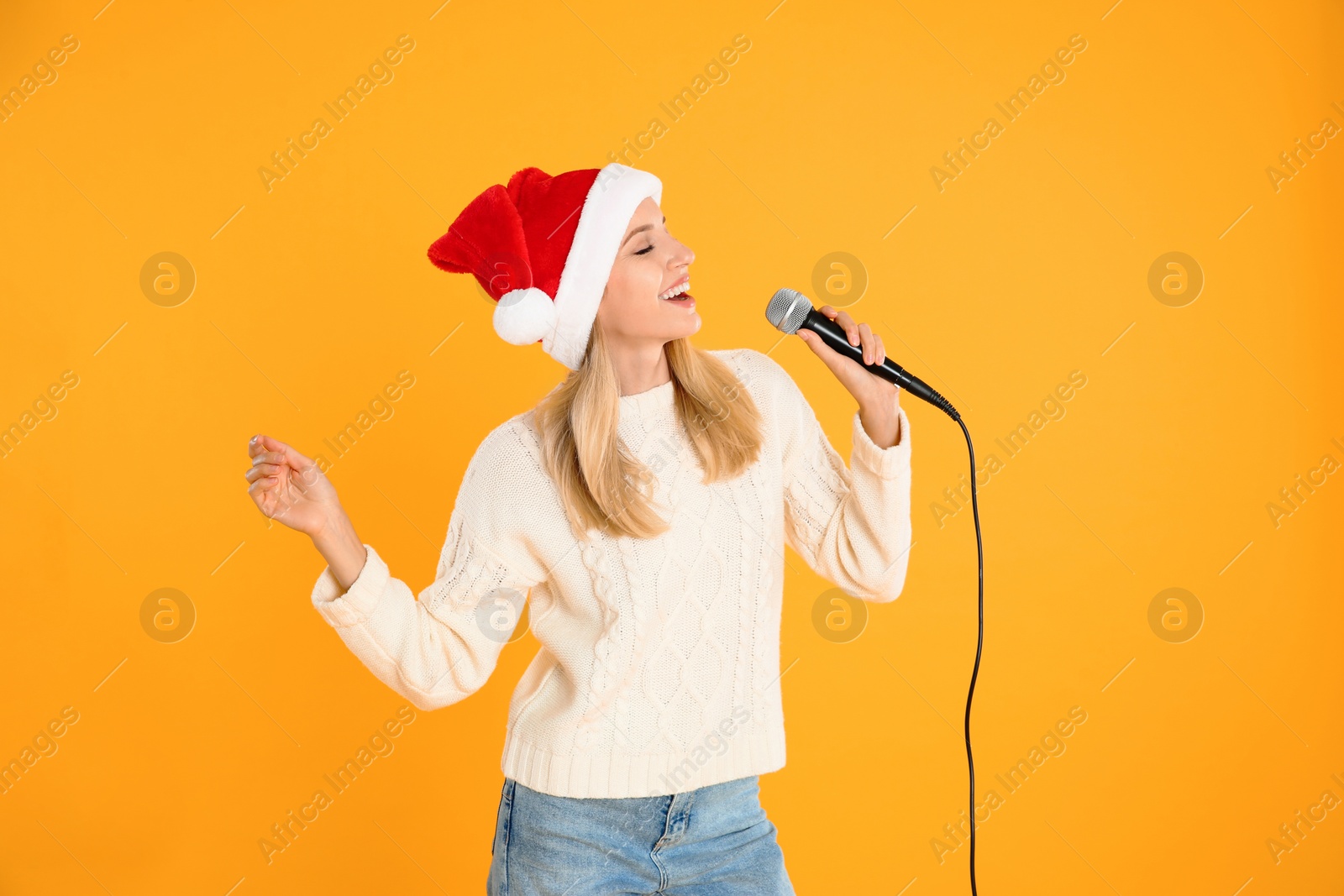 Photo of Emotional woman in Santa Claus hat singing with microphone on yellow background. Christmas music