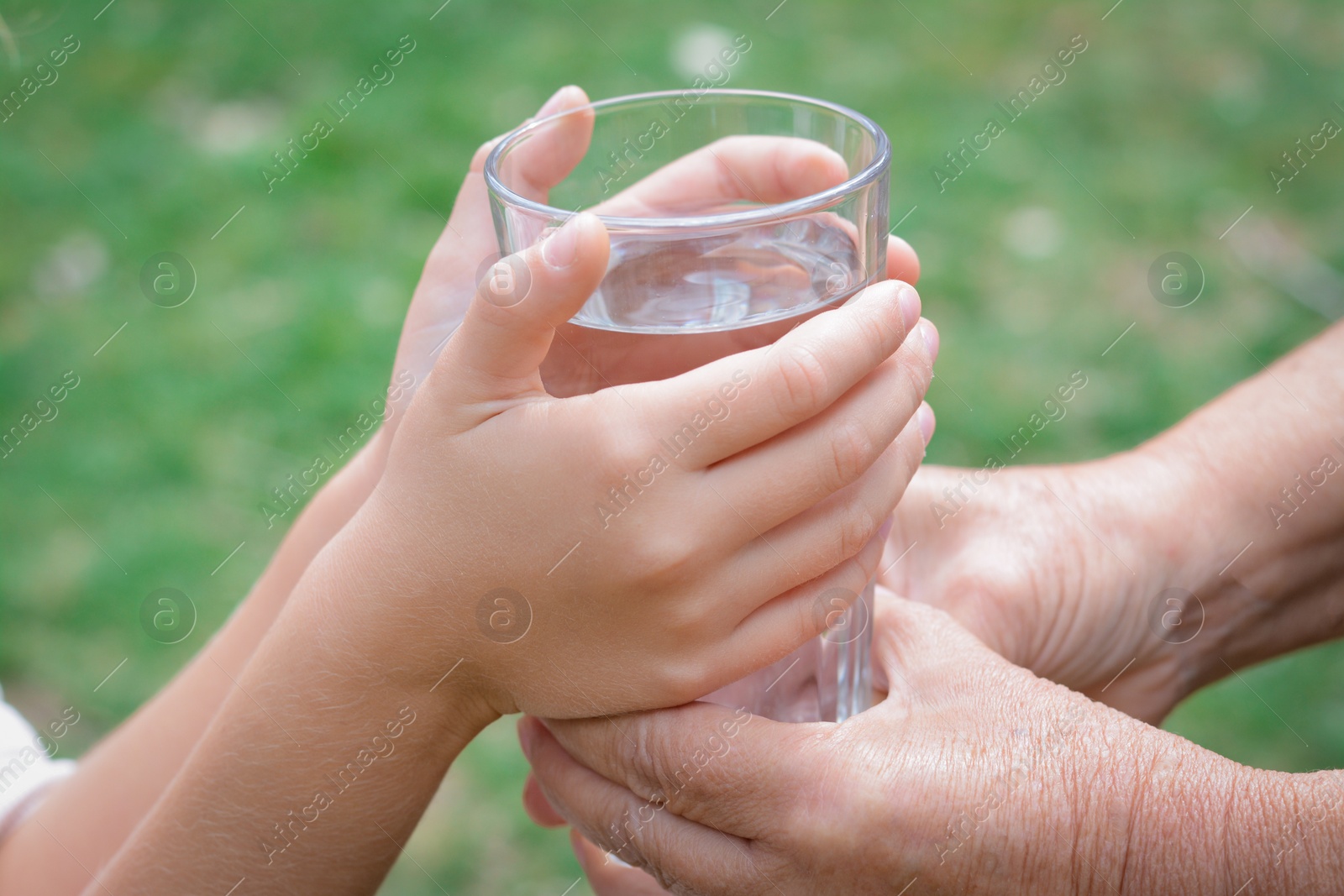 Photo of Child giving glass of water to elderly woman outdoors, closeup