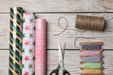 Photo of Different colorful wrapping paper rolls, scissors and threads on white wooden table, flat lay