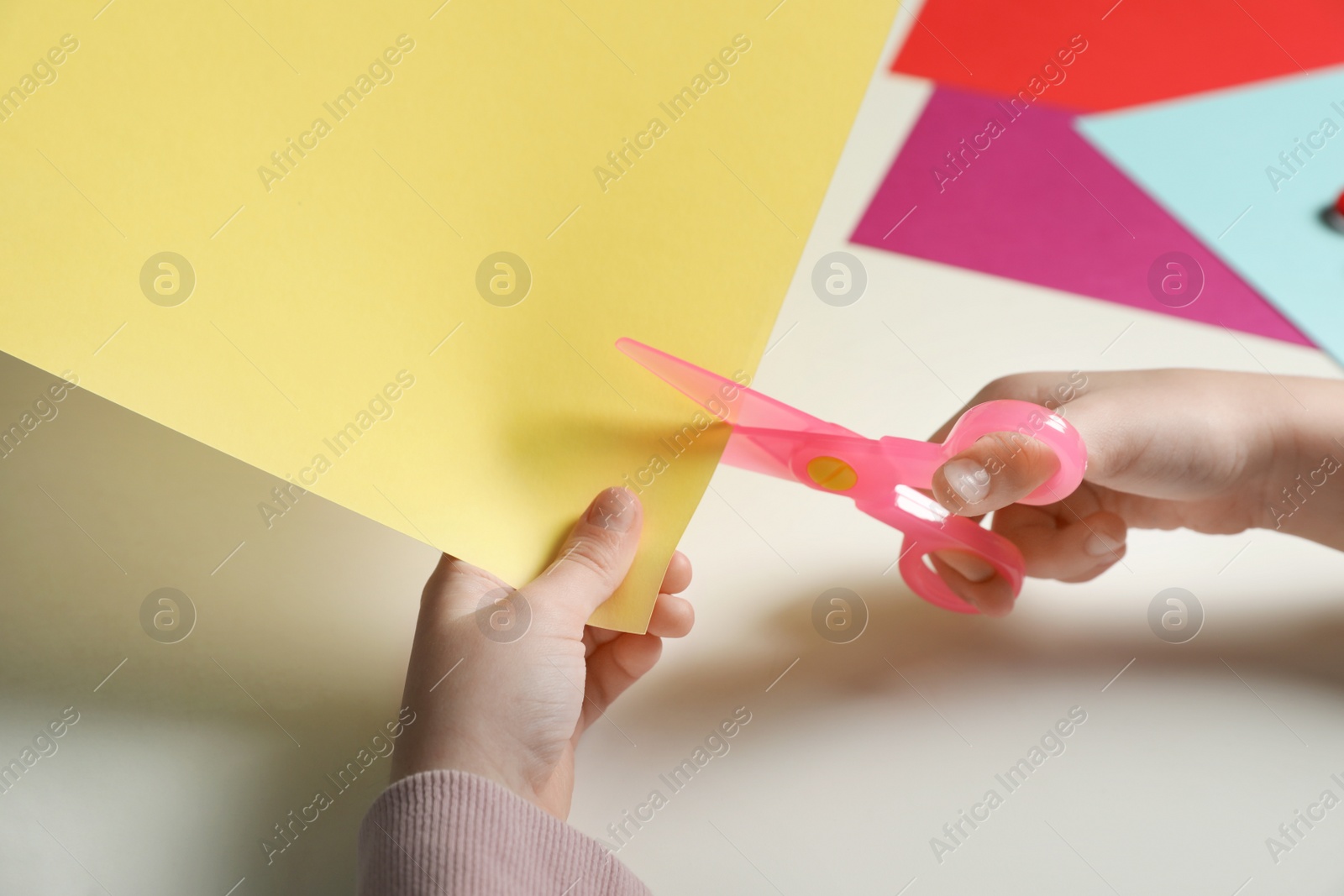 Photo of Child cutting color paper with plastic scissors at table, closeup