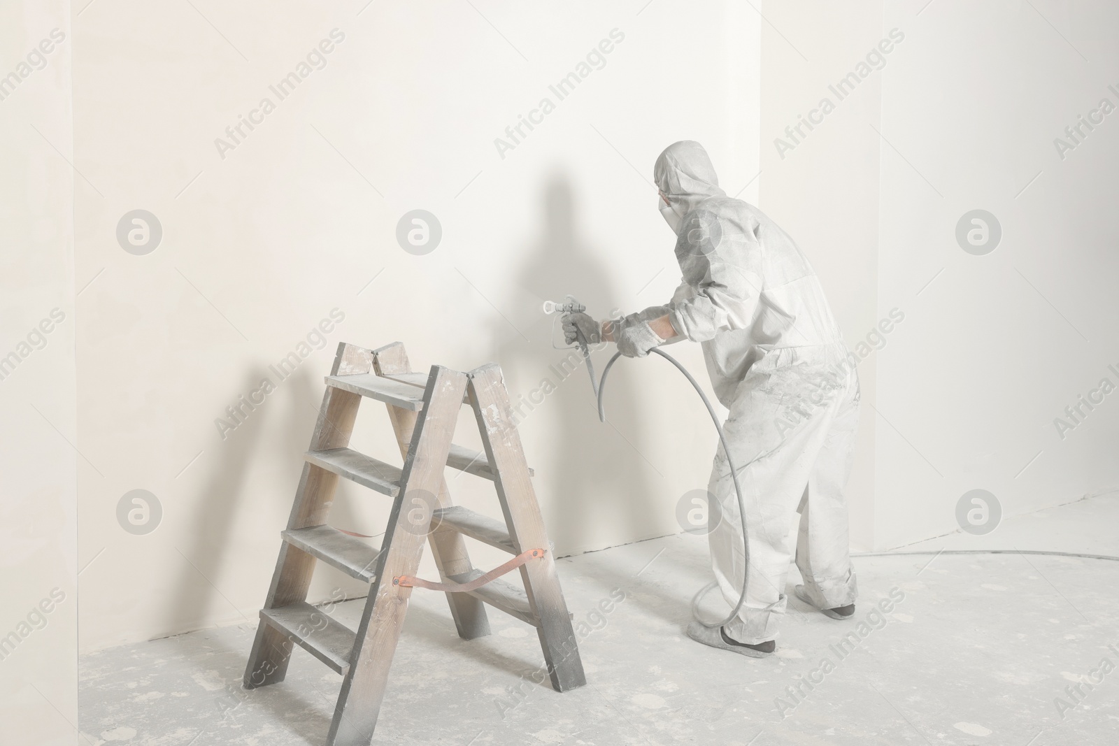 Photo of Decorator in protective overalls painting wall with spray gun indoors