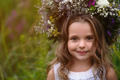 Cute little girl wearing wreath made of beautiful flowers in field, closeup