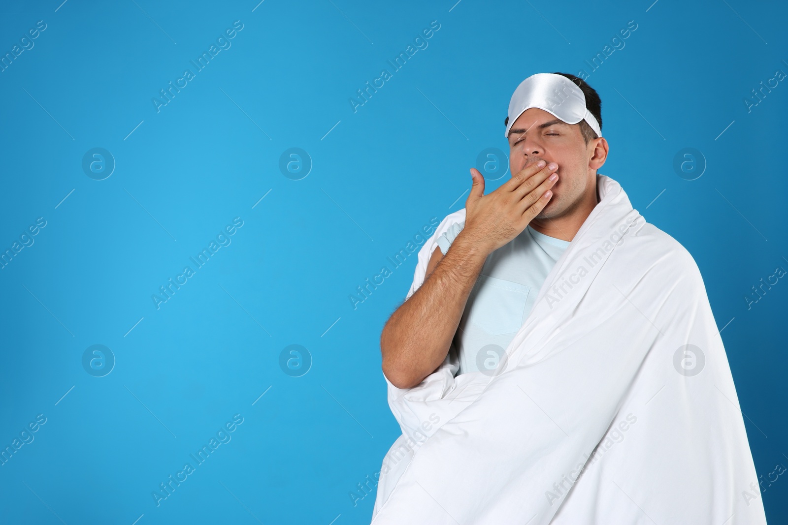 Photo of Man wrapped in blanket yawning on blue background. Space for text