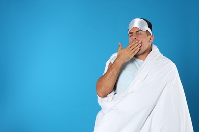 Man wrapped in blanket yawning on blue background. Space for text