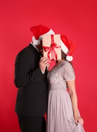 Photo of Couple in Santa hats covering their faces with Christmas gift on red background