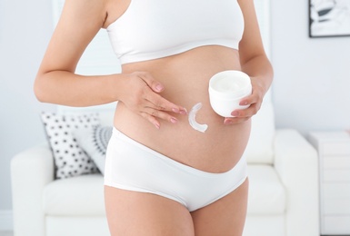 Pregnant woman applying body cream on belly at home, closeup