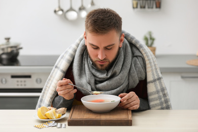 Sick young man eating tasty soup to cure flu at table in kitchen