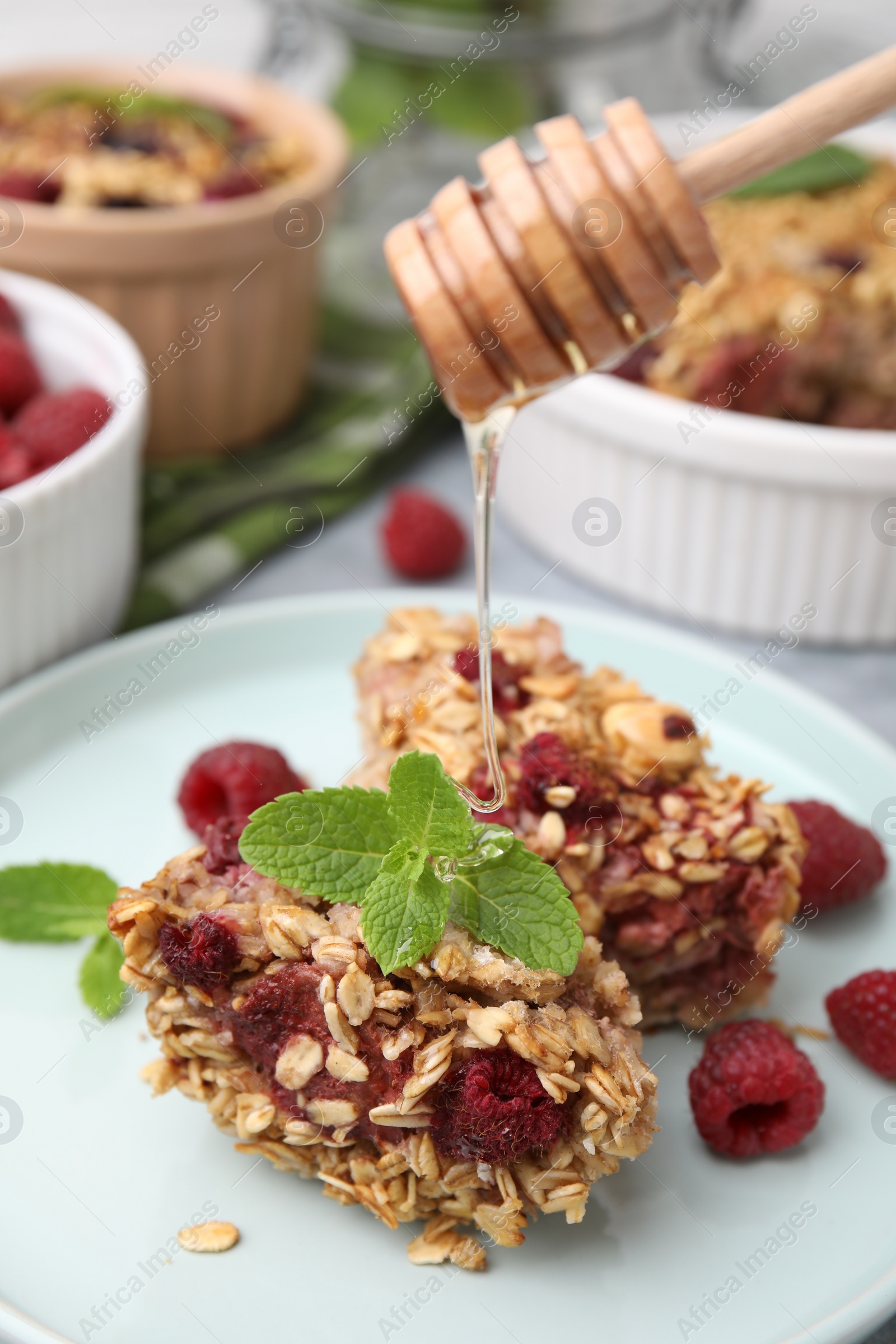 Photo of Pouring honey from dipper onto baked oatmeal with raspberries on table, closeup