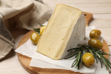 Photo of Board with piece of tasty camembert cheese, olives and rosemary on light wooden table, closeup