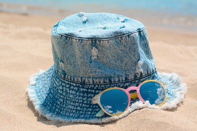 Jeans hat and sunglasses on sand, closeup. Beach accessories