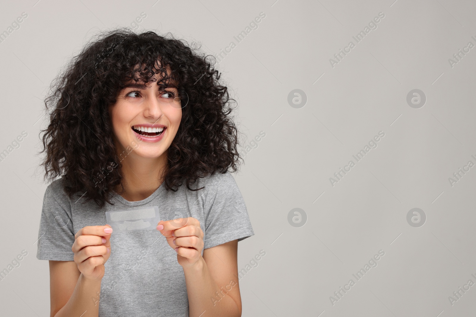 Photo of Young woman holding teeth whitening strips on light grey background, space for text