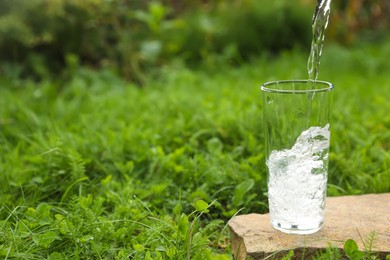 Photo of Pouring fresh water into glass on stone in green grass outdoors. Space for text