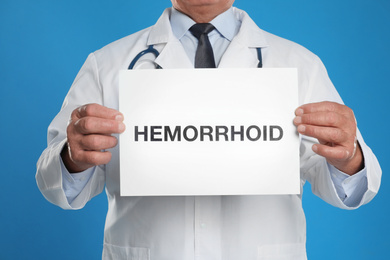 Photo of Doctor holding sign with word HEMORRHOID on blue background, closeup