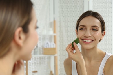 Photo of Young woman using aloe leaf to take care of skin near mirror indoors