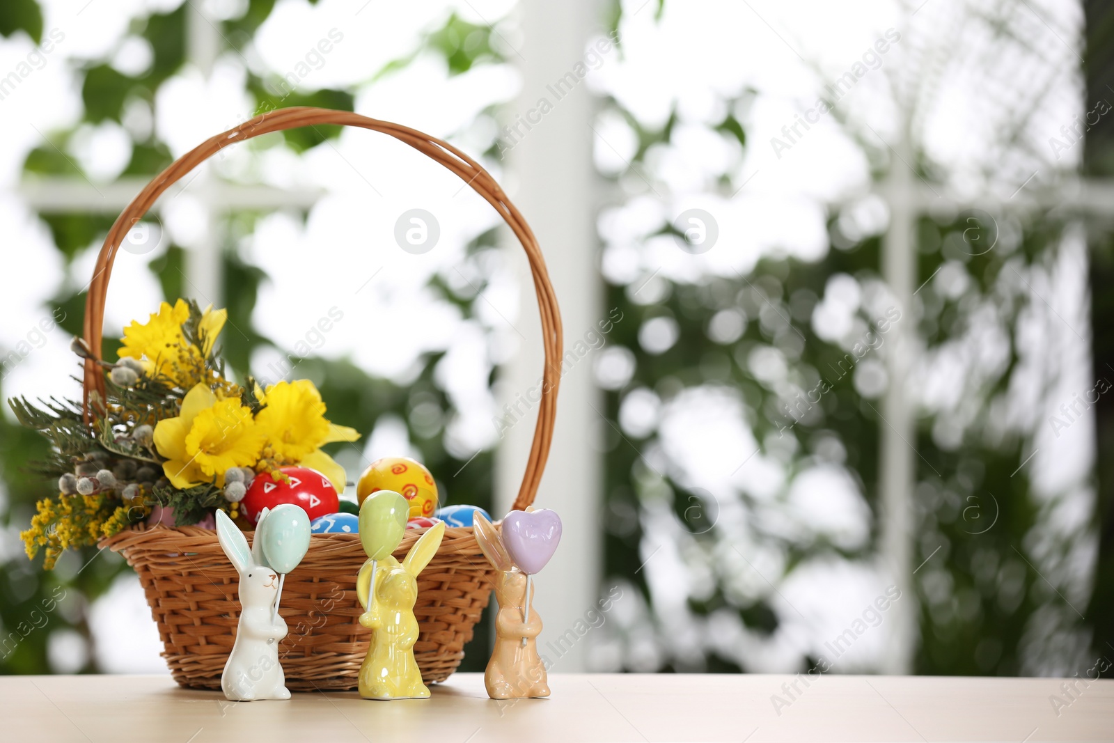 Photo of Festive composition with Easter eggs in wicker basket on table against blurred window, space for text