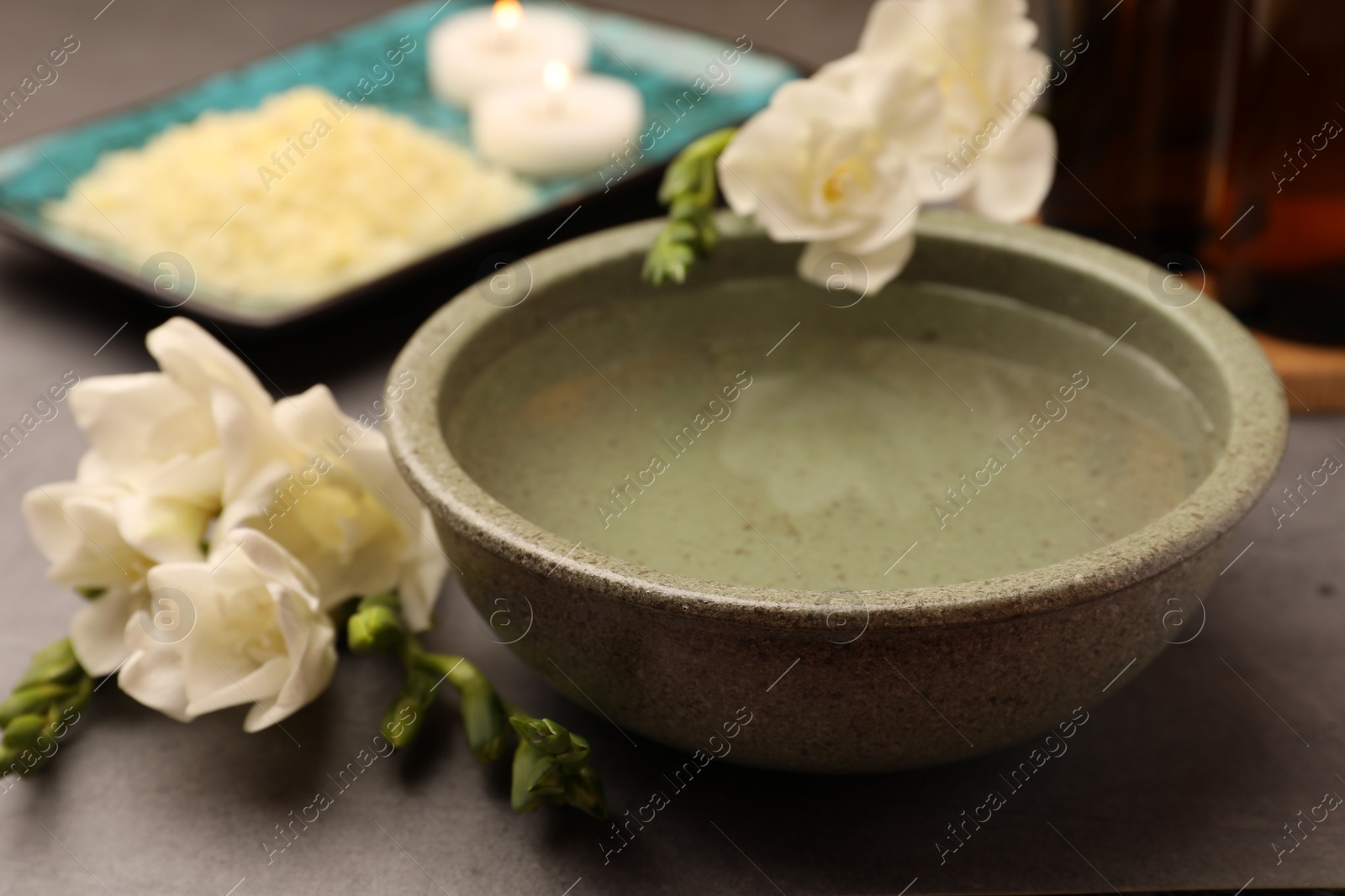 Photo of Bowl of essential oil and beautiful flowers on grey table, closeup. Aromatherapy treatment