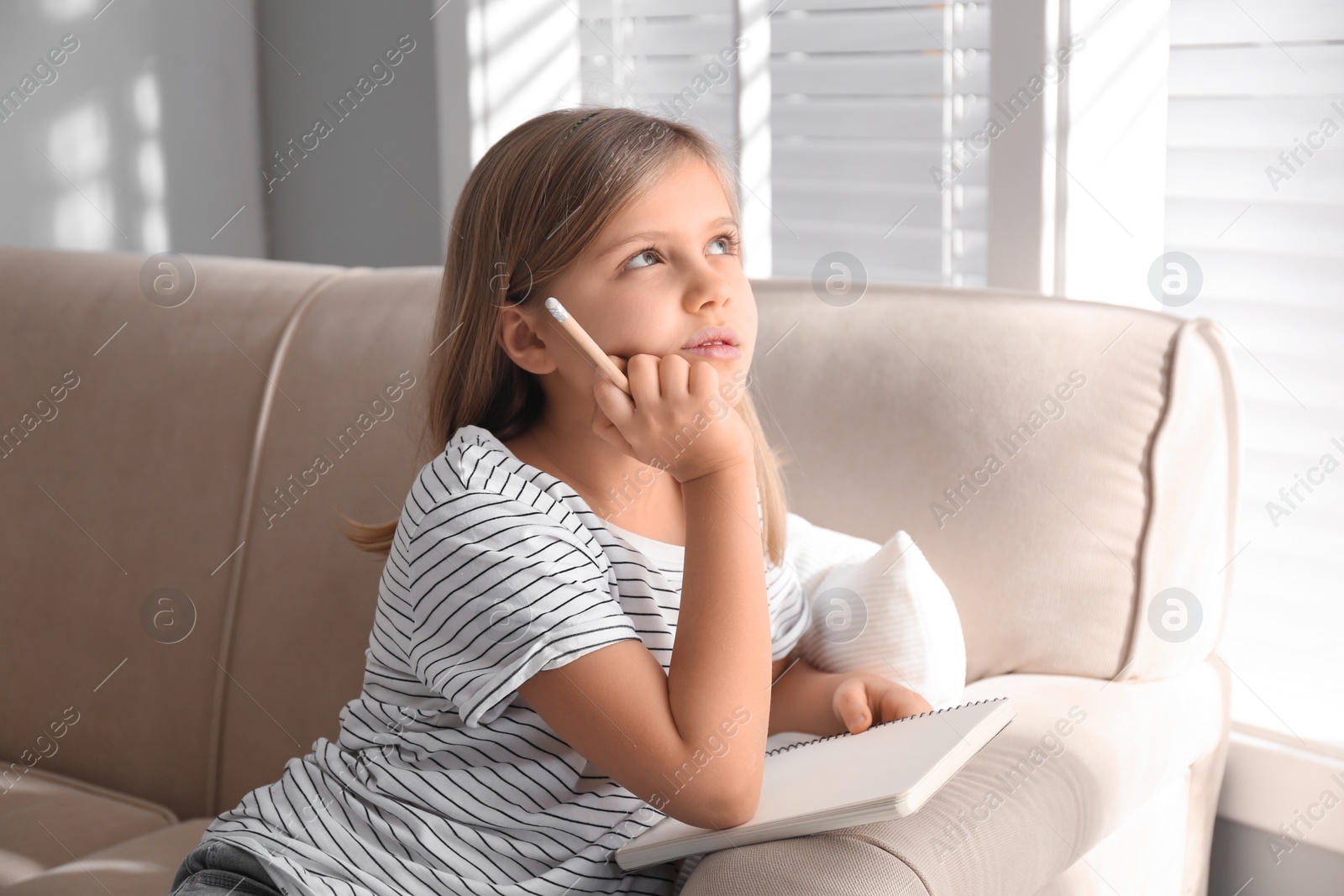 Photo of Thoughtful little girl with pencil and notebook on couch at home