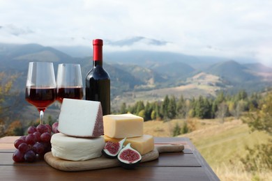 Photo of Different types of delicious cheeses, snacks and wine on wooden table against mountain landscape. Space for text