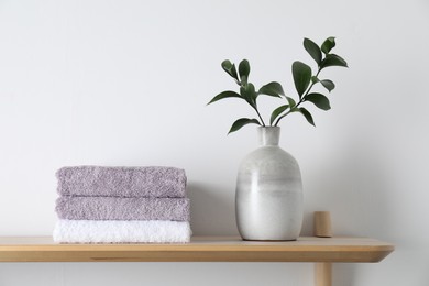 Stacked terry towels and green branches in vase on wooden shelf near white wall