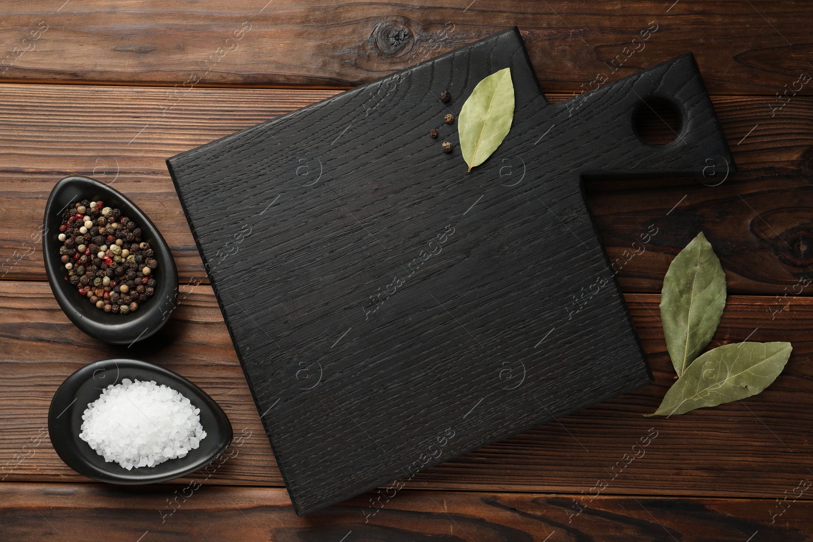 Photo of Black cutting board, salt, pepper and bay leaves on wooden table, flat lay. Space for text