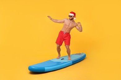 Photo of Happy man in Santa hat balancing on SUP board against orange background