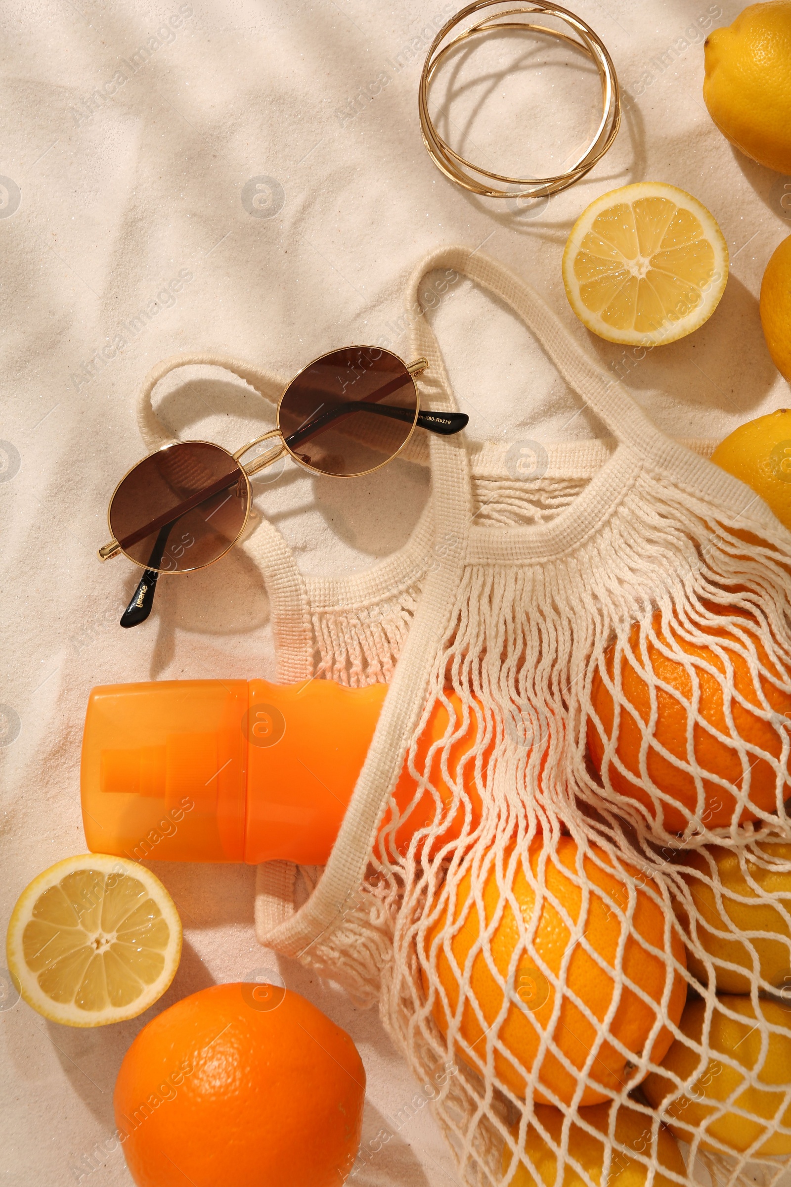 Photo of String bag with sunglasses, fruits and sunscreen on sand, flat lay