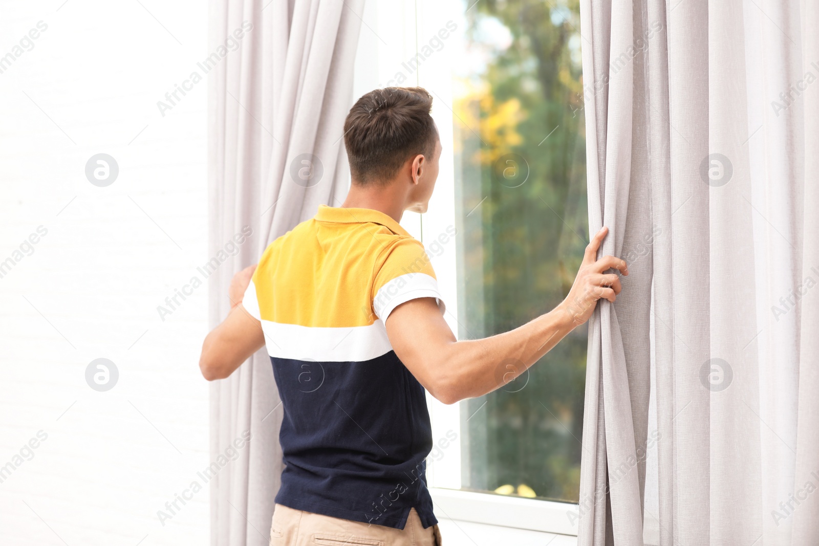 Photo of Young man opening window curtains at home