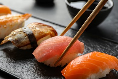 Photo of Taking delicious nigiri sushi with chopsticks from serving board on black table, closeup