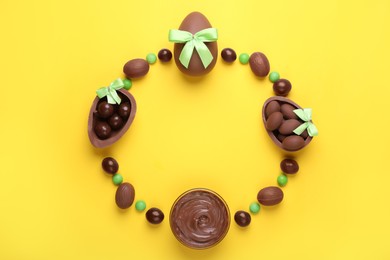 Photo of Frame made of tasty chocolate eggs with light green bows, paste and candies on yellow background, flat lay. Space for text