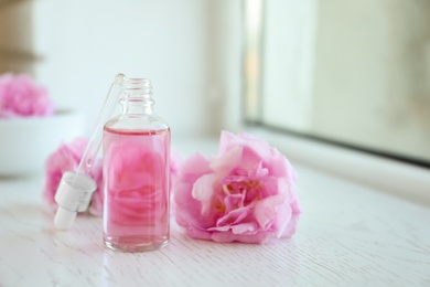 Photo of Bottle of rose essential oil, pipette and fresh flowers on window sill, space for text