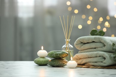 Photo of Spa composition. Towels, stones, reed air freshener and burning candles on white marble table against blurred lights, space for text