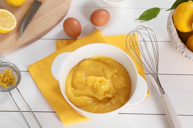 Delicious lemon curd in bowl, ingredients, whisk and sieve on white wooden table, above view