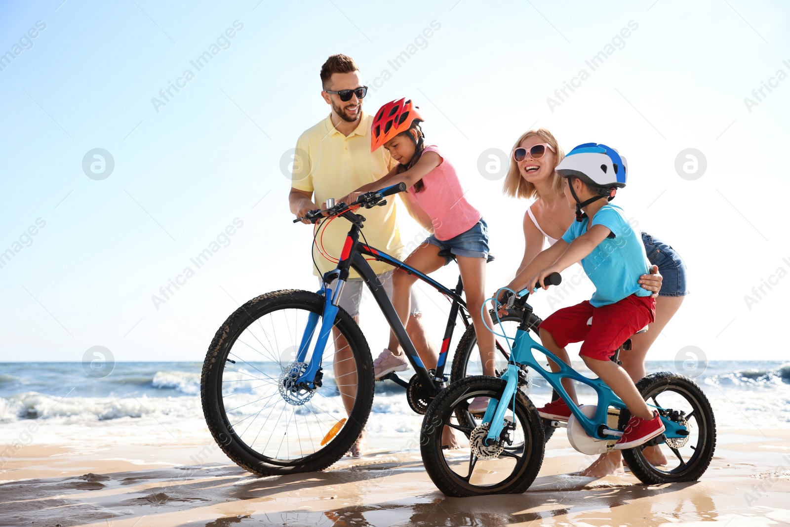 Photo of Happy parents teaching children to ride bicycles on sandy beach near sea