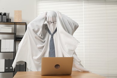 Photo of Overworked ghost. Man covered with white sheet using laptop at wooden table in office