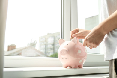 Photo of Woman putting money into piggy bank at window sill indoors, closeup. Space for text