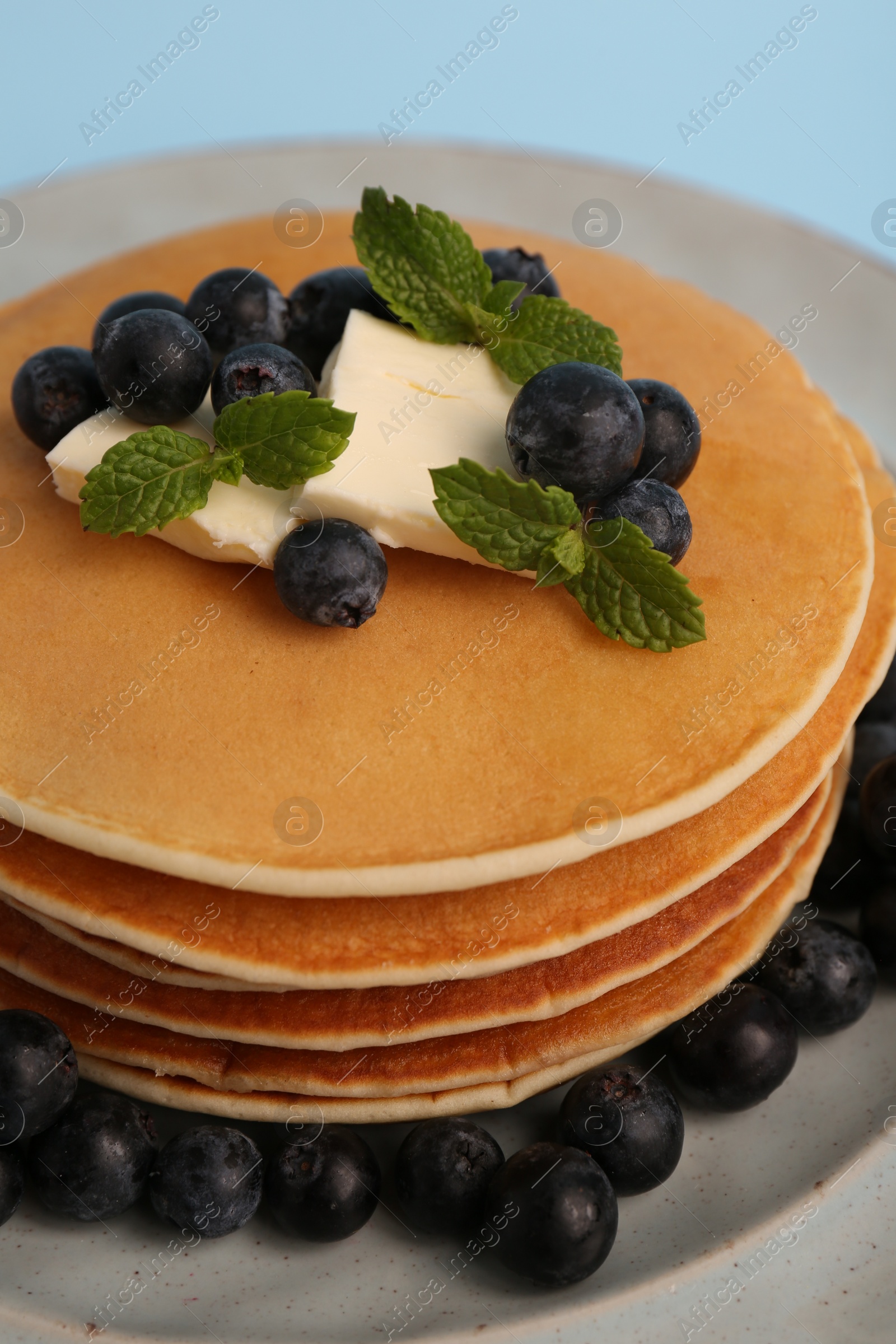 Photo of Stack of tasty pancakes with blueberries, butter and mint on light blue background, closeup