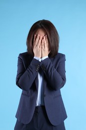 Upset woman in suit closing her face with hands on light blue background