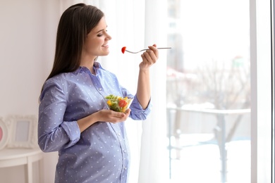 Photo of Young pregnant woman eating vegetable salad near window at home