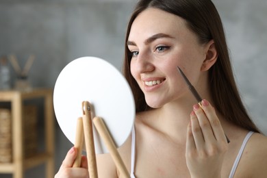 Smiling woman drawing freckles with pen in front of little mirror indoors