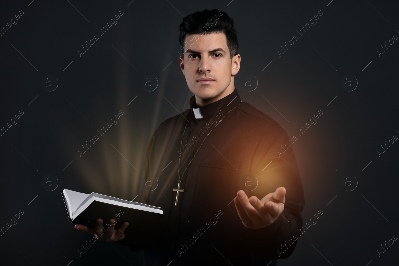 Image of Priest in cassock holding Bible with holy light on black background