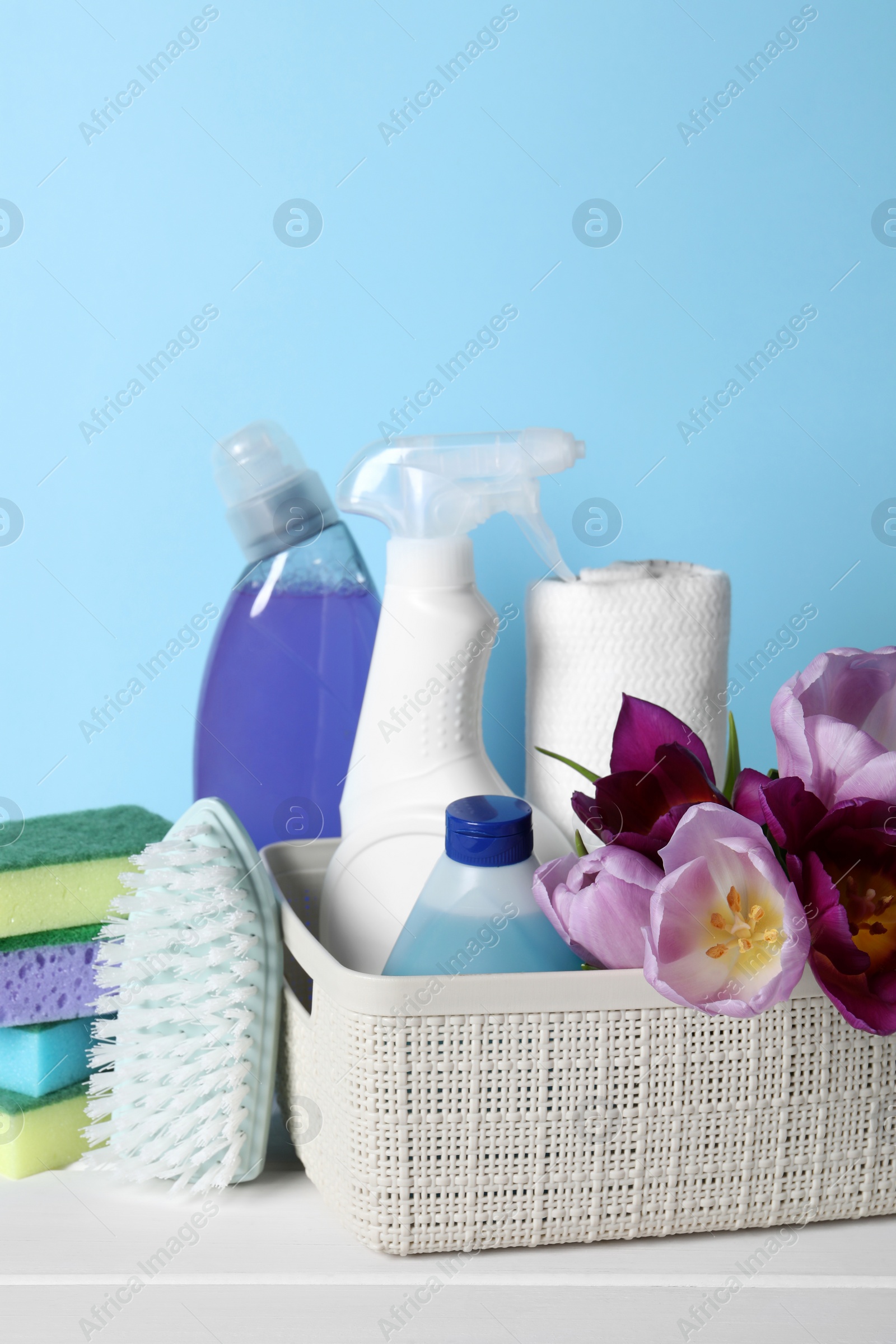 Photo of Spring cleaning. Basket with detergents, flowers and tools on white wooden table against light blue background