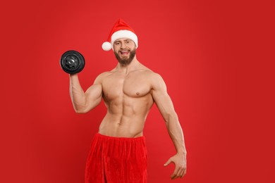 Muscular young man in Santa hat with dumbbell on red background