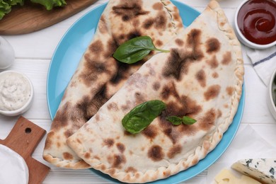 Delicious cheese calzones, basil and sauces on white wooden table, flat lay